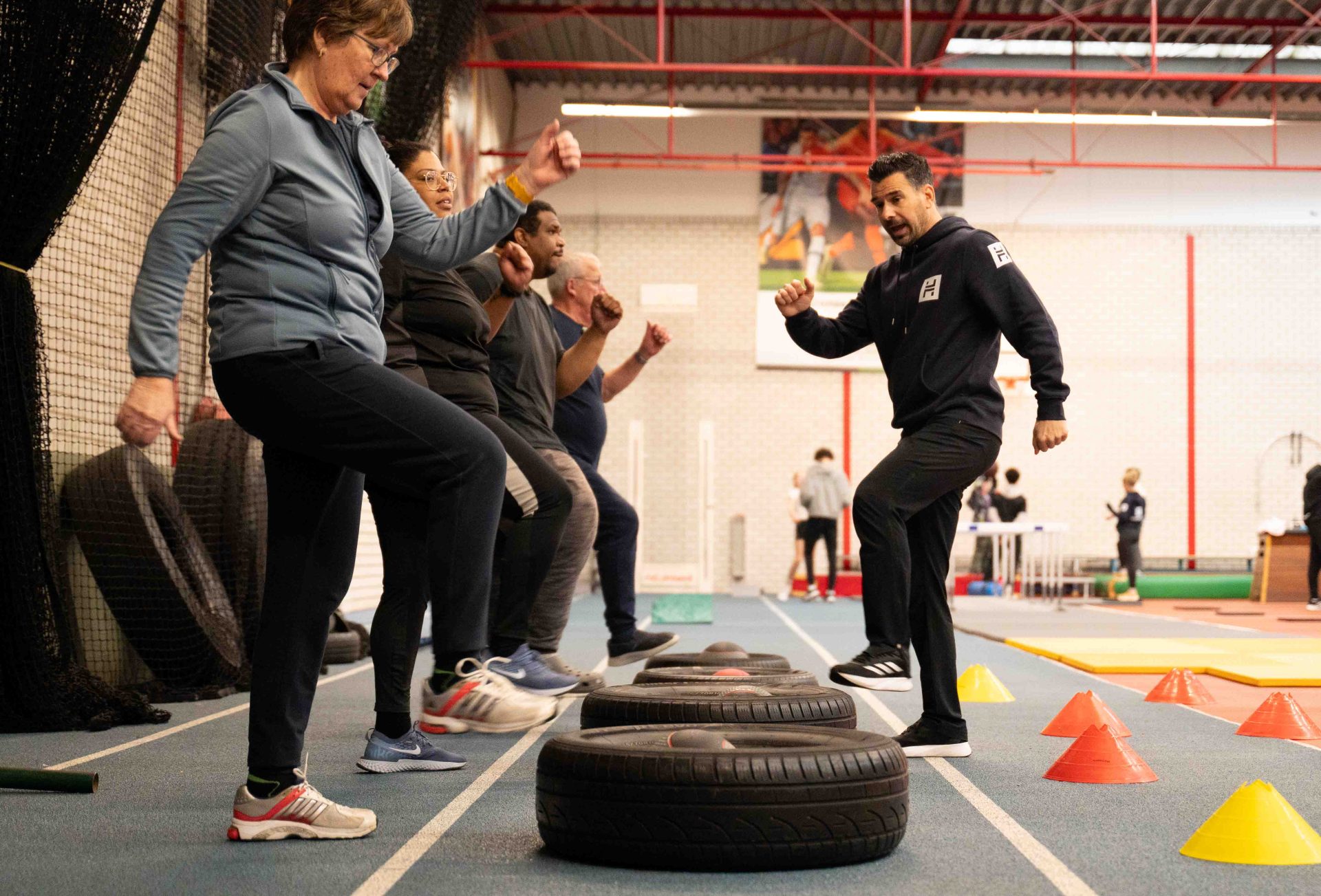Groep deelnemers doet high knees oefening over autobanden onder begeleiding van personal trainer in sporthal
