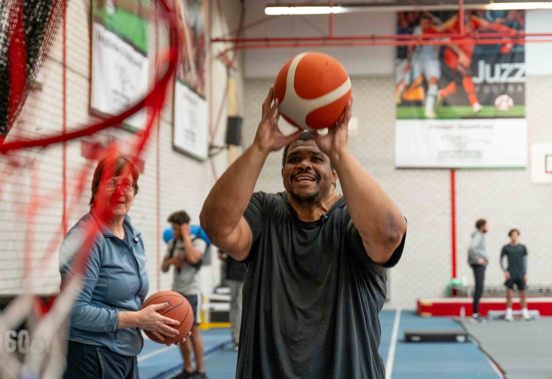 Man schiet basketbal richting basket in sporthal, met oudere vrouw op de achtergrond die ook een basketbal vasthoudt