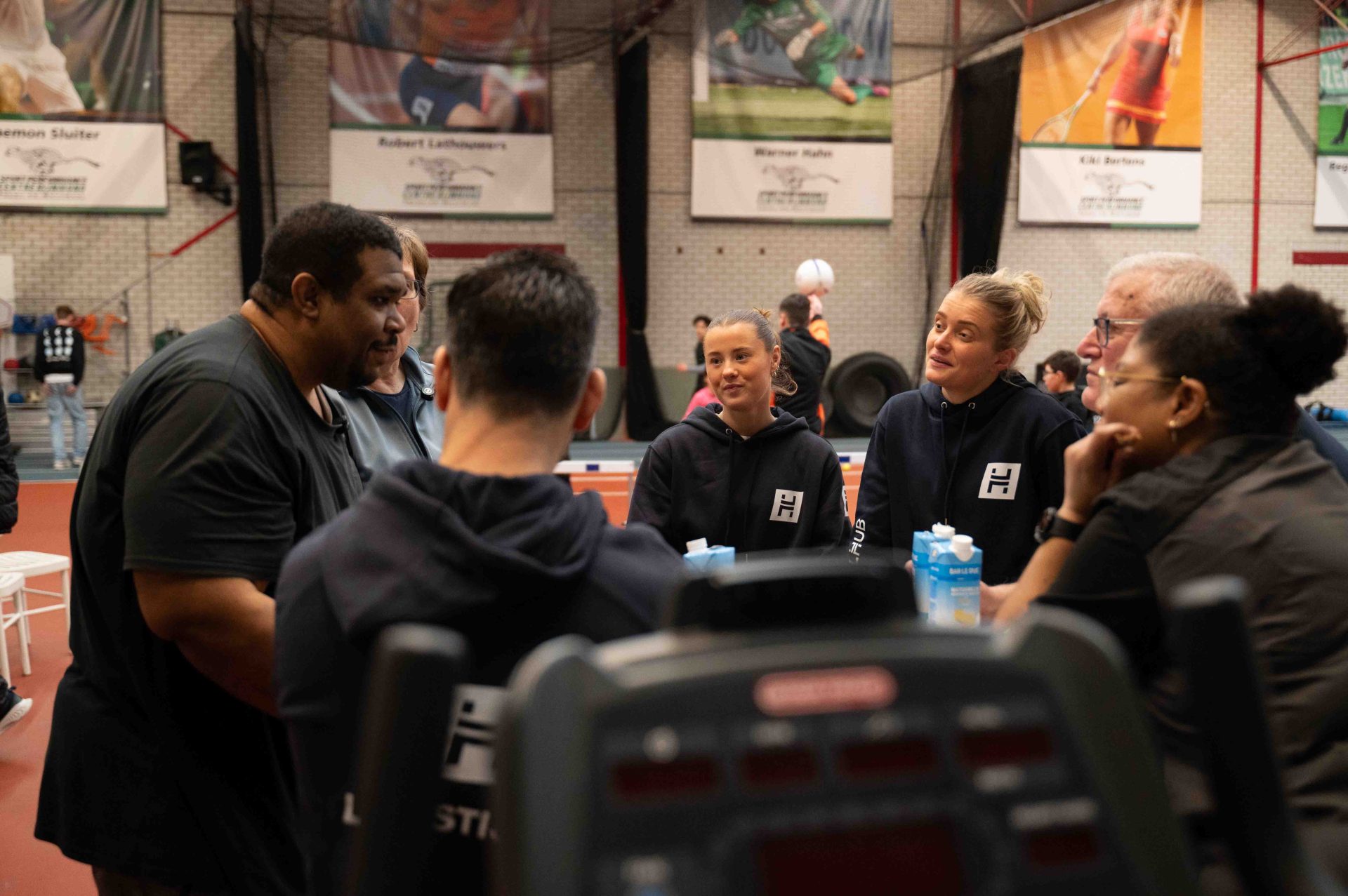 Groep deelnemers en coaches in gesprek tijdens teammoment in de sportschool
